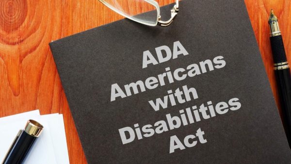 Black folder labeled 'ADA Americans with Disabilities Act' on a wooden desk, with glasses, a pen, and white papers nearby.