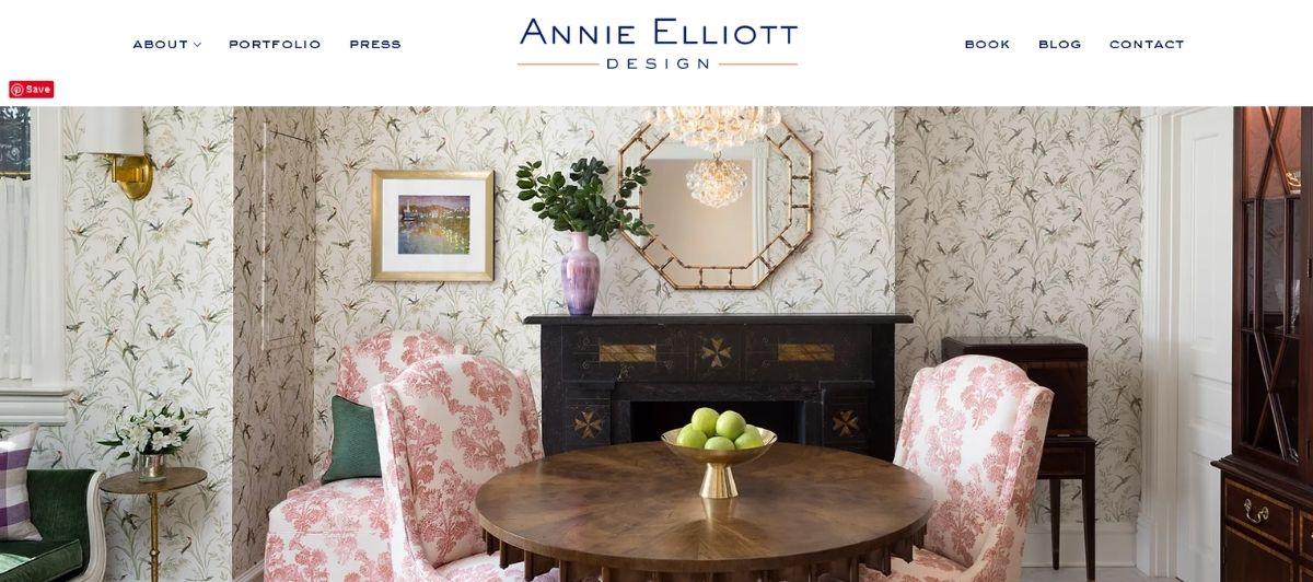 Elegant living room interior: round wooden table, pink floral chairs, and a dark mantle against patterned wallpaper with a chandelier above.