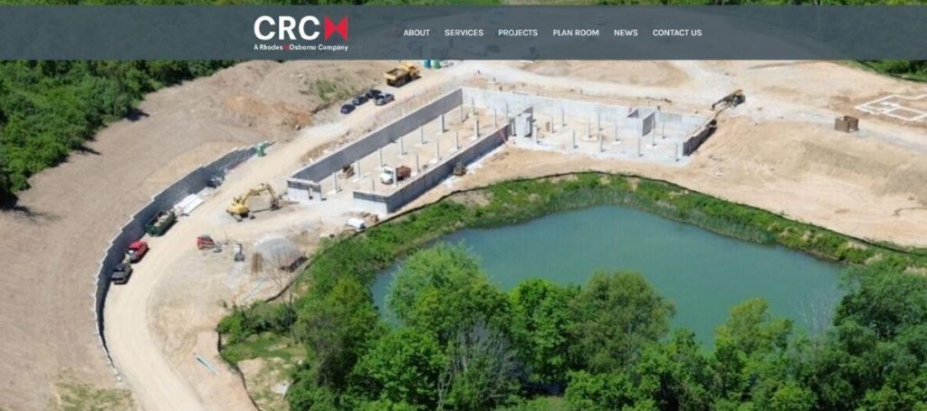 Aerial view of a construction site with a partially built rectangular structure beside a turquoise pond, surrounded by dirt roads and trees, with construction equipment on site.