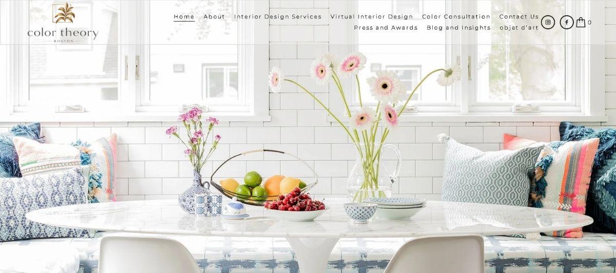 Bright dining area with a white marble table, colorful cushions, and vases of pink daisies and fruit centerpiece.