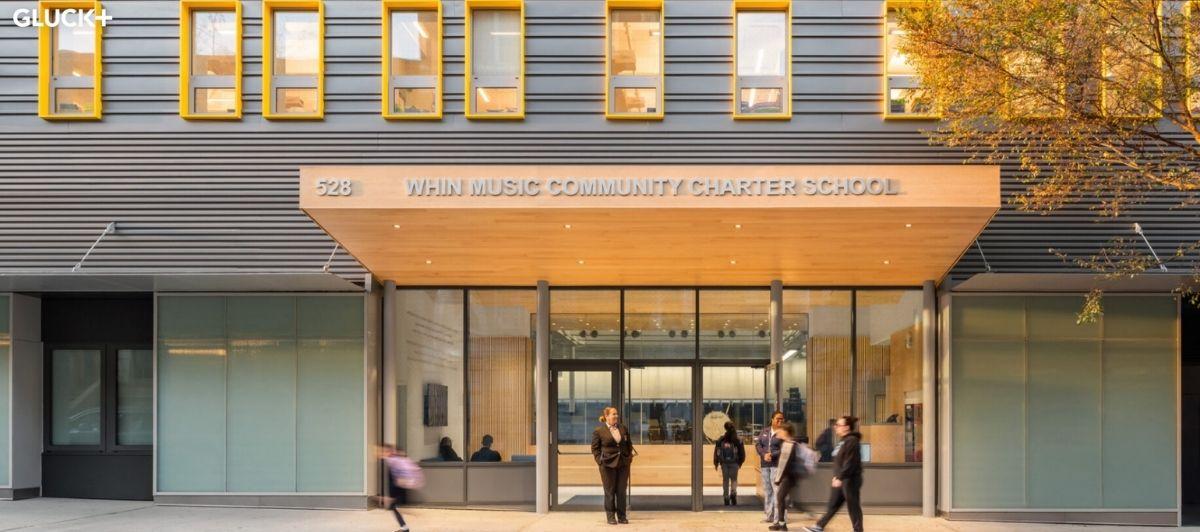 Facade of Whin Music Community Charter School with a wooden entry canopy, yellow-framed windows, and people entering the building.