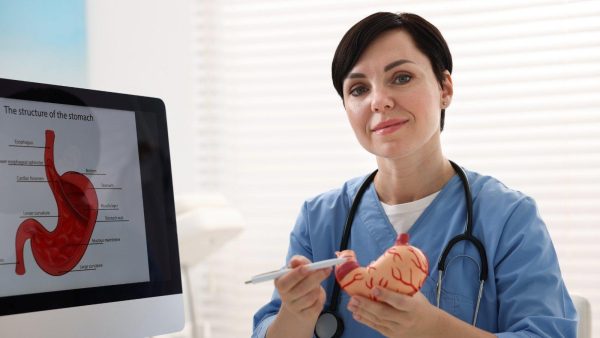 Female healthcare professional in blue scrubs explains stomach anatomy using a 3D model while a diagram of the stomach appears on a computer screen behind her
