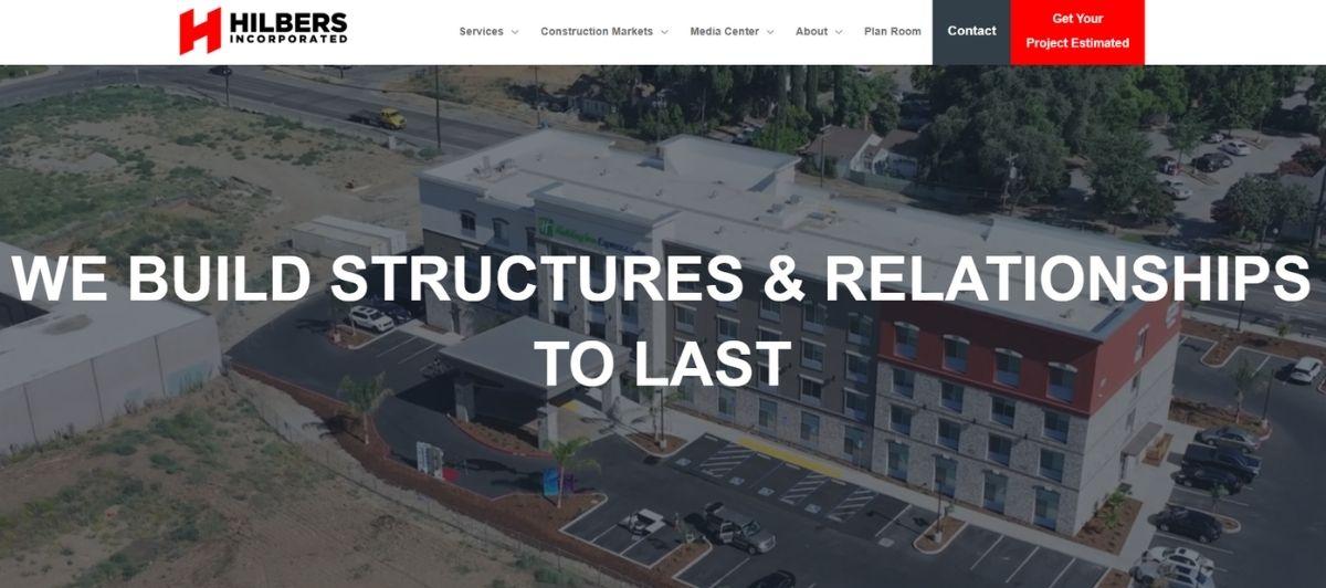 Aerial view of a modern building complex with a parking lot and trees, with the slogan 'WE BUILD STRUCTURES & RELATIONSHIPS TO LAST' overlaid.