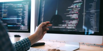 Software developer pointing at code on a large monitor with multiple screens in a bright office.