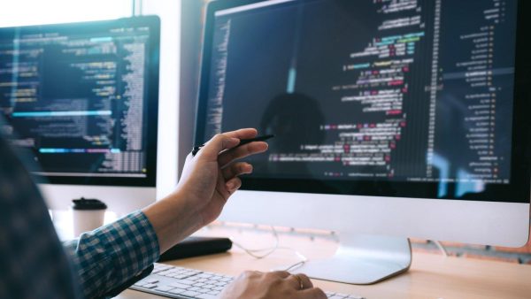 Software developer pointing at code on a large monitor with multiple screens in a bright office.