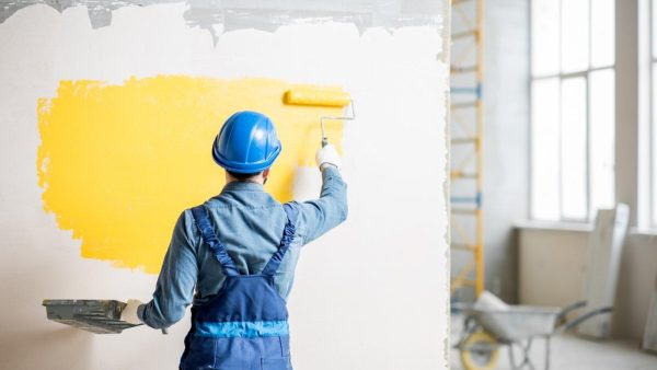 Construction worker in a blue hard hat and overalls rolling bright yellow paint on an interior wall.