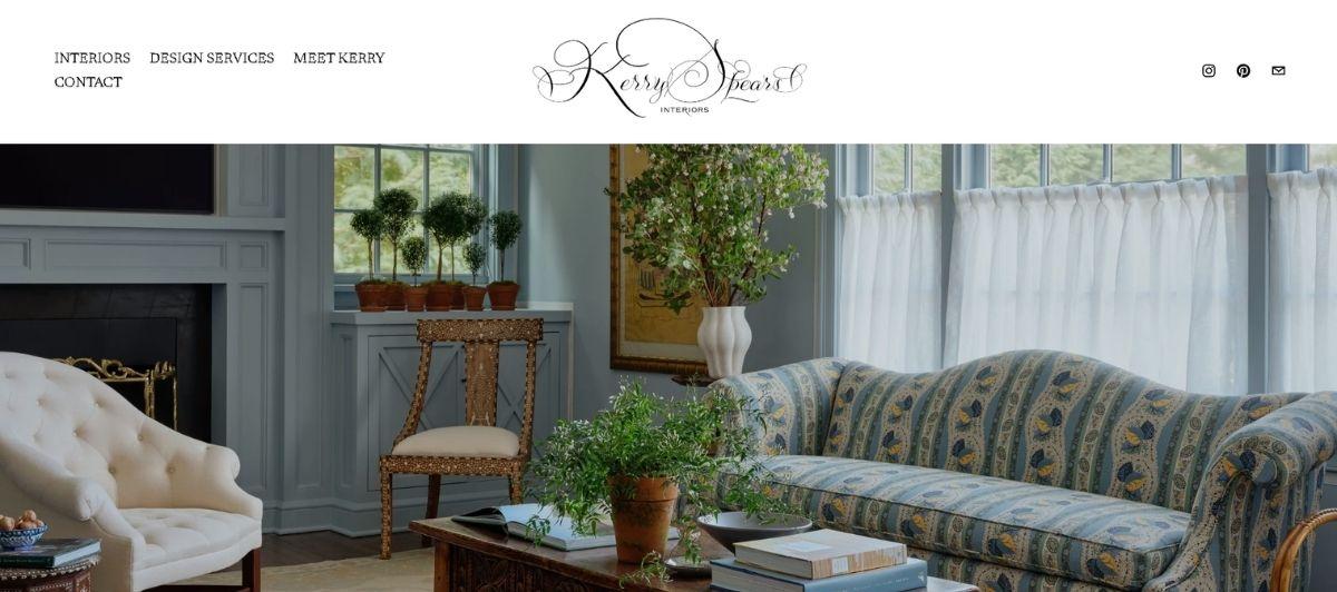 Elegant living room with a tufted cream chair, carved wooden chair, potted plants, and a patterned blue sofa by a large window.