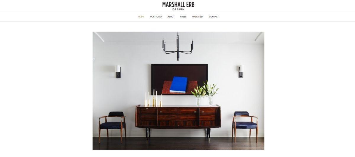Mid-century wood sideboard with candles and a vase of flowers, flanked by blue chairs in a bright living room, with a wall-mounted artwork above and a black chandelier overhead.