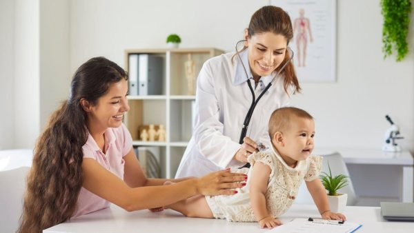 Pediatrician uses a stethoscope to examine a baby on an examination table while a smiling mother-supporting caregiver watches nearby.