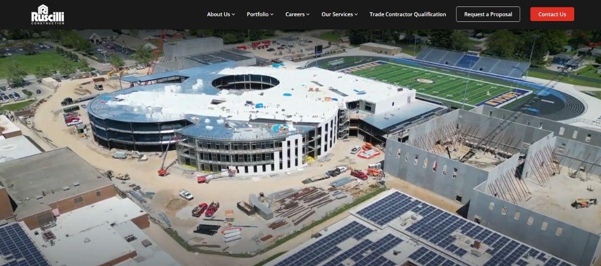 Aerial view of a large sports stadium under construction, with a circular centerpiece and surrounding buildings, trucks and cranes nearby.