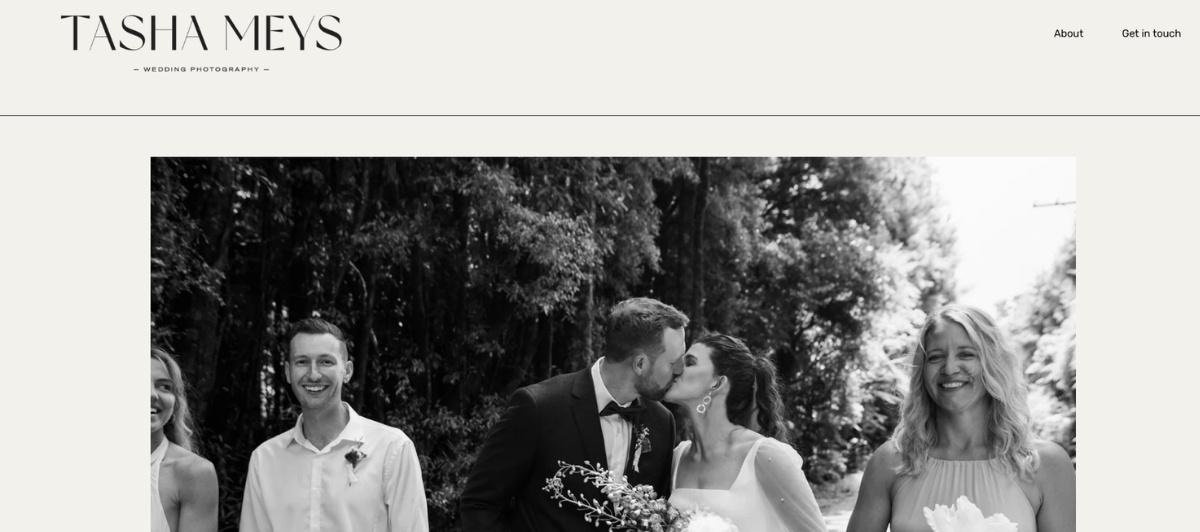 Bride and groom share a kiss at their wedding, surrounded by laughing guests outdoors in black and white.