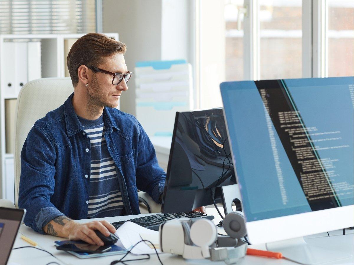 Man with glasses coding at a bright desk with two monitors showing lines of code, in a modern office.