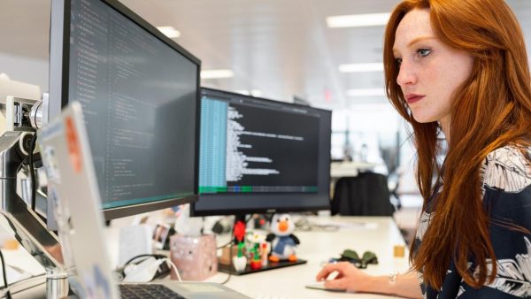 Red-haired woman coding at a workstation with two monitor screens displaying code in a bright office.