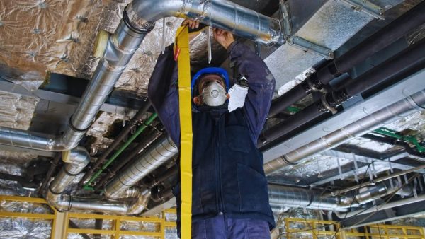 Maintenance worker in a hard hat and respirator tightens a yellow strap under overhead ductwork in an industrial space.