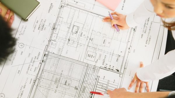 Architects review architectural blueprints on a table, one person pointing with a purple pencil to a floor plan area.