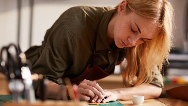 Woman in a craft workshop leans over a worktable, cutting material with a craft knife on a green cutting mat wearing a brown apron.