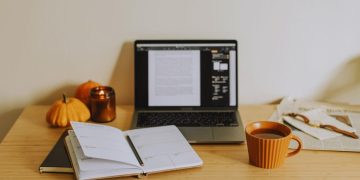 Autumn desk setup: laptop with an open planner, pumpkins, a candle, and an orange mug on a wooden table with a newspaper nearby