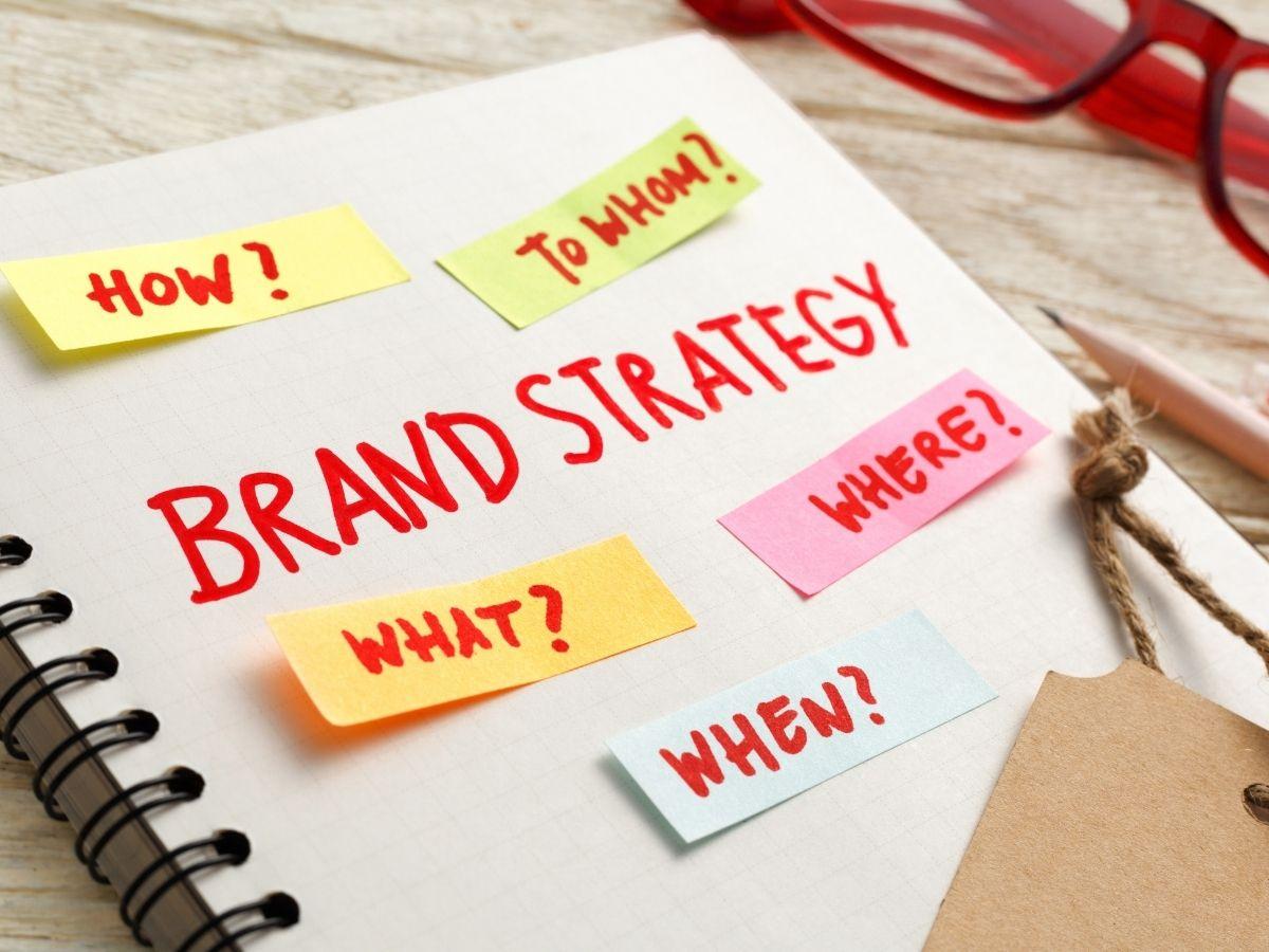 Notebook page labeled 'Brand Strategy' with colorful sticky notes asking How? To whom? What? Where? When? (bright red glasses in background)
