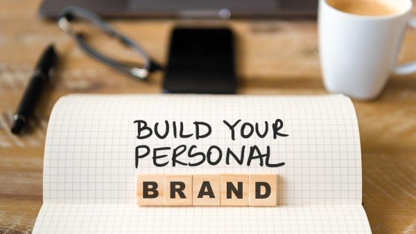 Notebook page with the message 'BUILD YOUR PERSONAL BRAND' and wooden tiles spelling BRAND, on a desk with a coffee mug in the background.