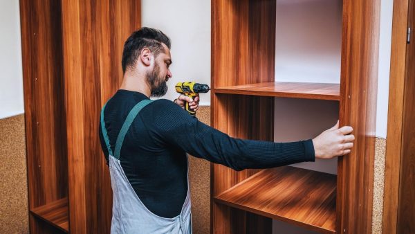 Bearded man in a black shirt and gray overalls drills a wooden cabinet shelf with a yellow cordless drill.