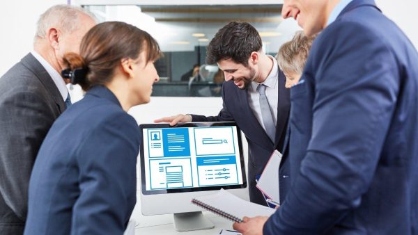 Team of professionals gathered around a desktop computer, reviewing blue UI layout mockups on screen in a meeting center.