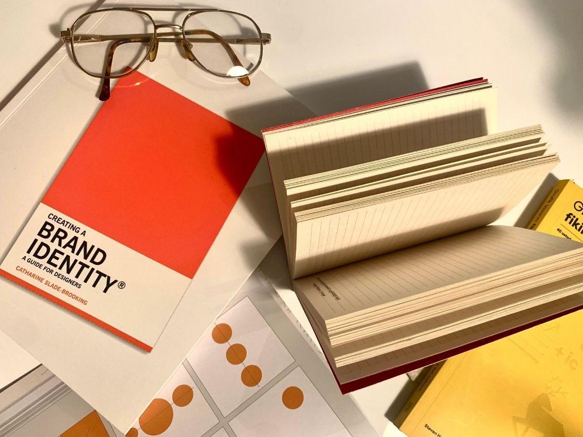 Open notebook with cream pages and an orange-covered design book titled 'Creating a Brand Identity' beside gold-rimmed glasses on a white desk.