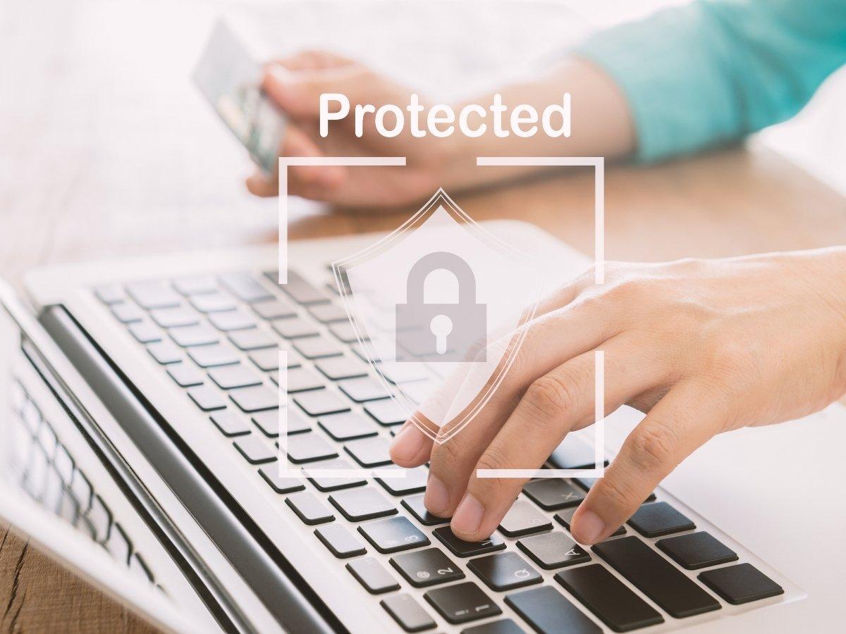 Close-up of hands typing on a laptop with a translucent shield and padlock icon labeled 'Protected' (cybersecurity illustration)