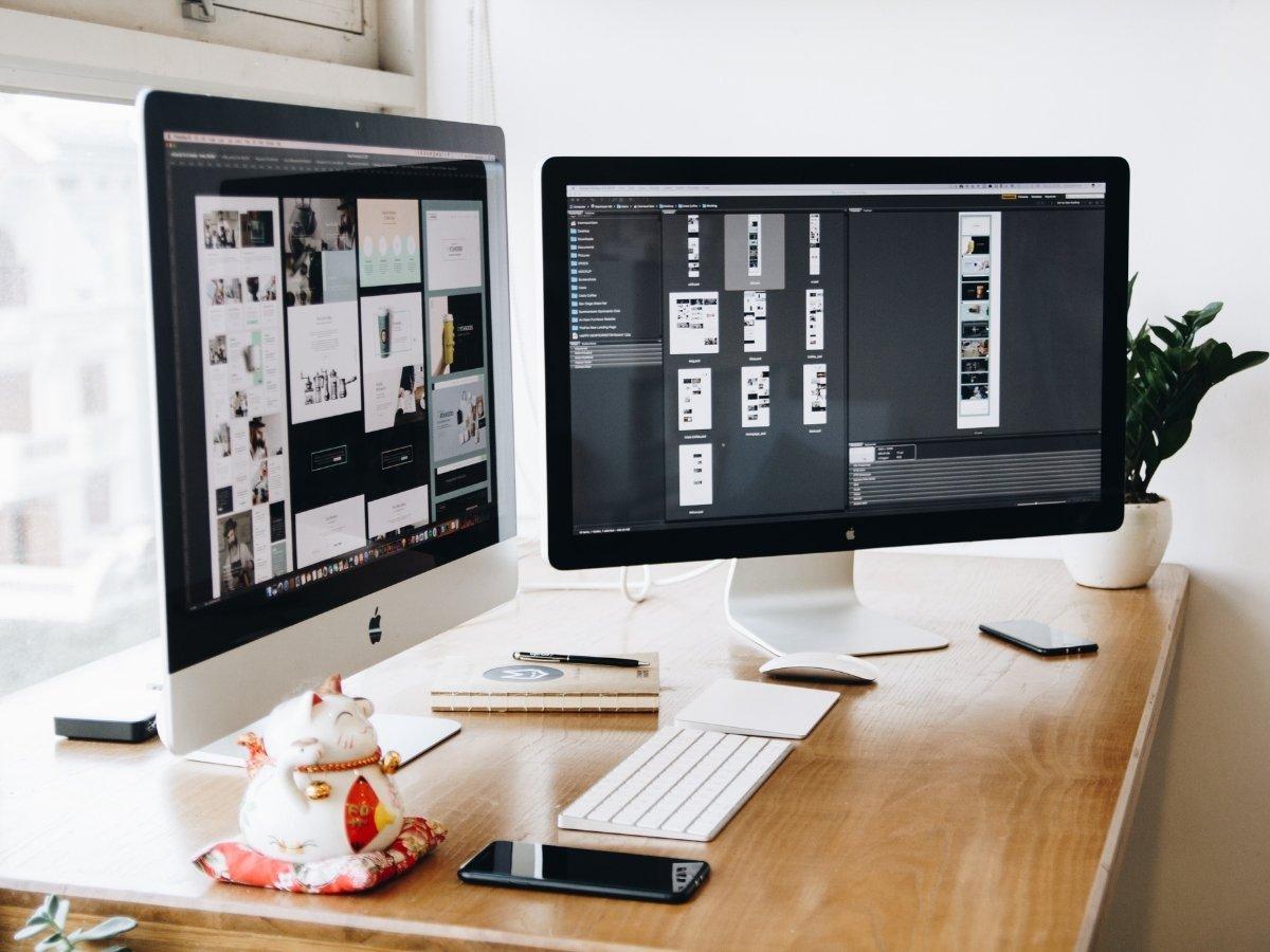 Two iMacs on a wooden desk show design work, with a keyboard, mouse, notebook, plant, and smartphone nearby.