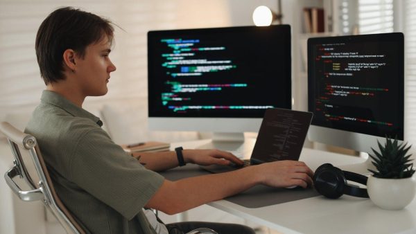 Side profile of a person coding at a white desk with two monitors showing colorful code.