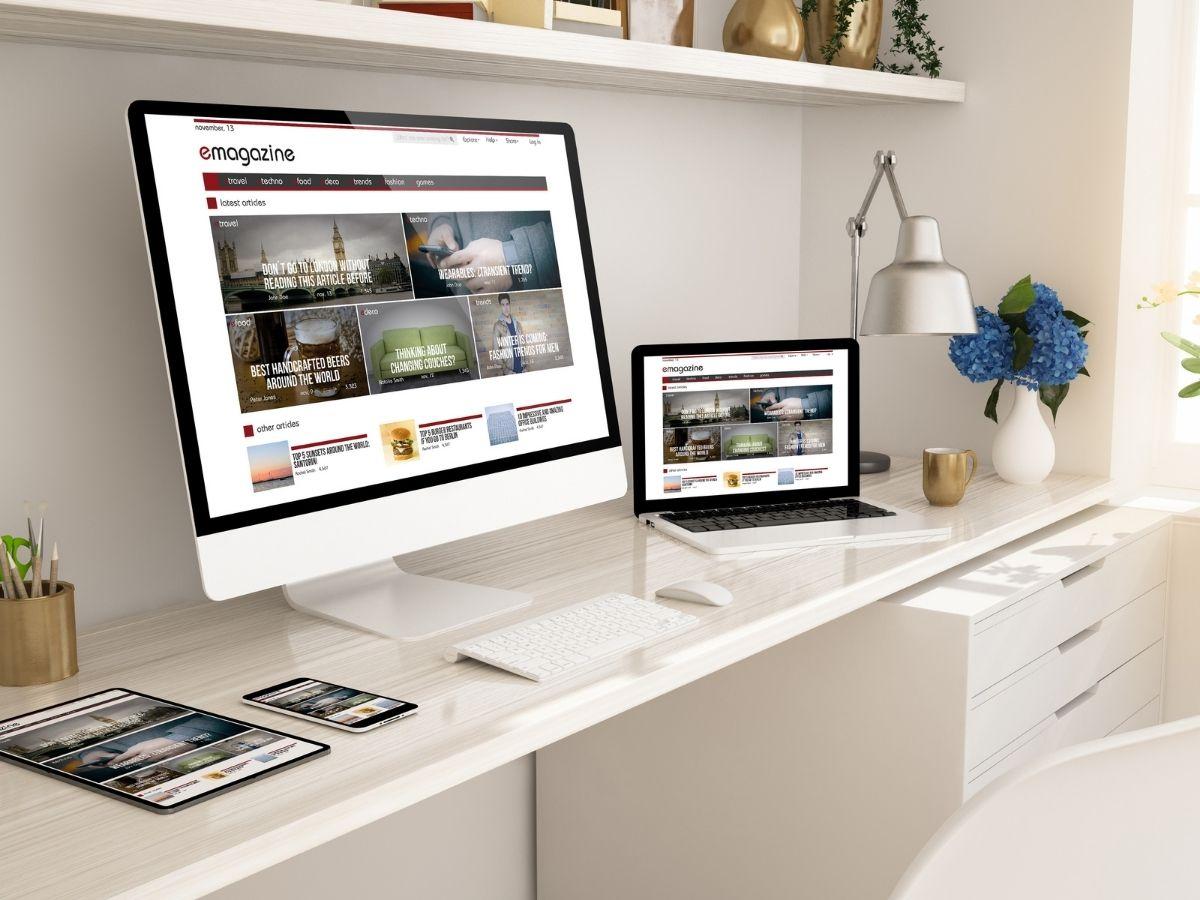 White desk setup with a large monitor, a laptop, tablet, and a phone all displaying the emagazine homepage on multiple devices.