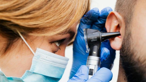 Healthcare professional wearing a mask and blue gloves uses an otoscope to examine a patient's ear, close-up view.