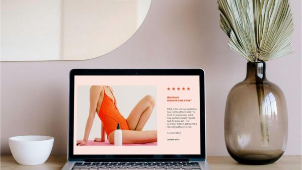 Laptop screen shows an ad: a woman in a red swimsuit posing on a pink background with a sunscreen bottle, text on the right; desk has a white bowl and a tall brown vase with dried leaves nearby.