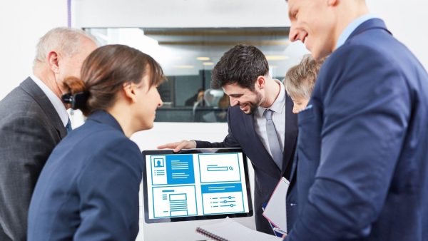 Team of professionals gathered around a tablet showing blue UI layouts during a meeting.
