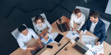 Overhead view of four professionals around a table examining charts and graphs on paper and laptops.