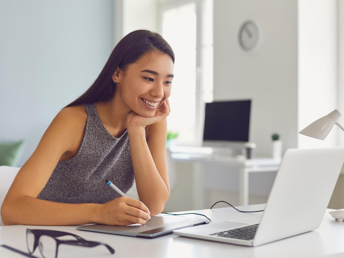 Woman smiling while drawing on a graphic tablet at a bright white desk with a laptop.