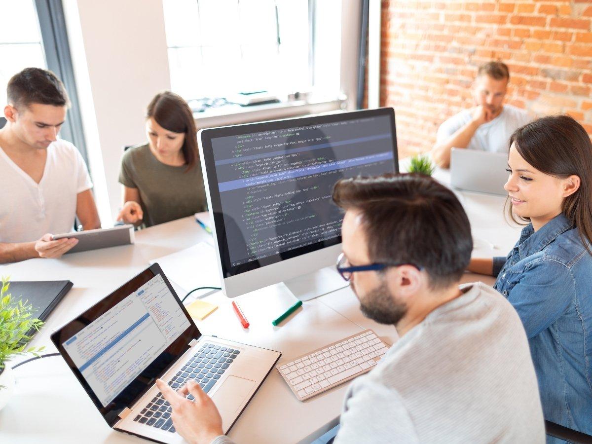 Team of five developers in a bright office, one man coding on a desktop monitor while others work on laptops near a brick wall.