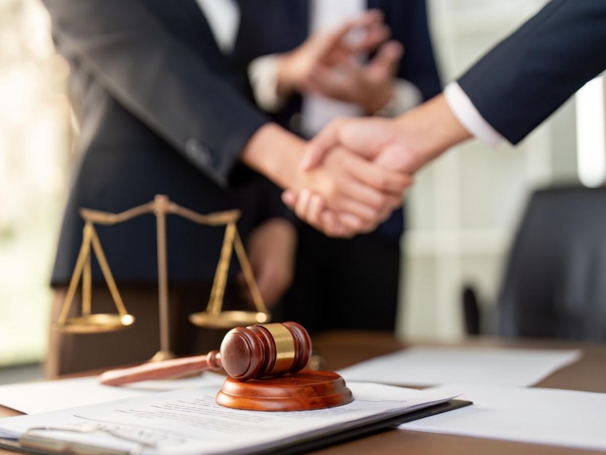 Gavel on a desk with documents and scales of justice, while two professionals shake hands in the blurred background.