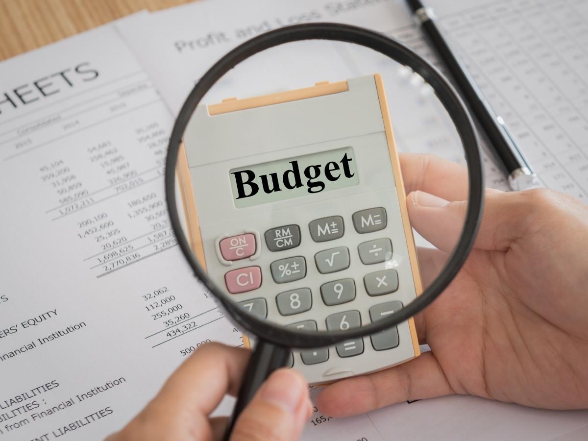 Close-up of a hand using a magnifying glass over a calculator displaying 'Budget' with financial sheets in the background.