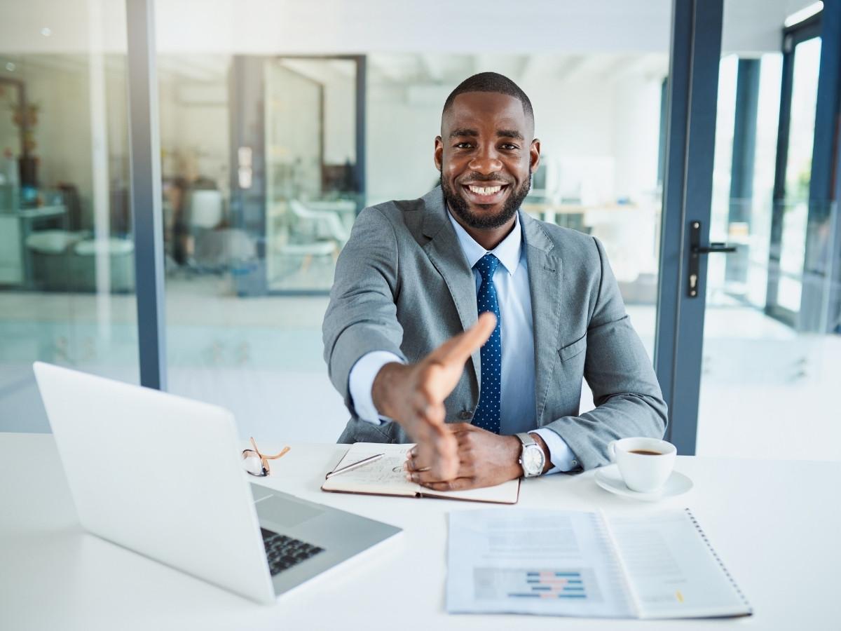Professional man in a gray suit extends a handshake across a desk in a bright office.