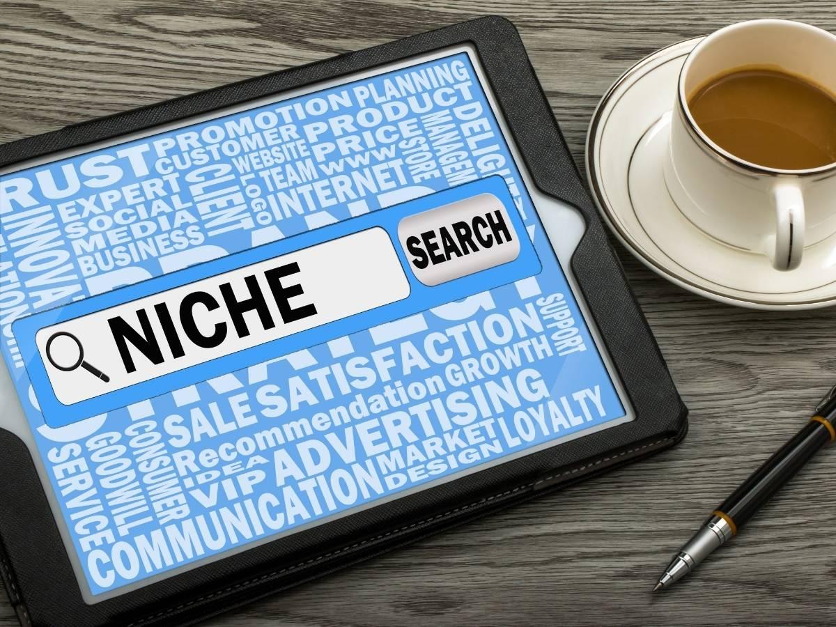 Tablet screen with a large NICHE search bar over a blue word-cloud background; a coffee cup and pen on the desk nearby.