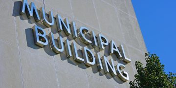 Exterior wall of a municipal building with large metal letters spelling MUNICIPAL BUILDING, blue sky to the right and a tree in the foreground.