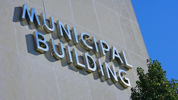 Exterior wall of a municipal building with large metal letters spelling MUNICIPAL BUILDING, blue sky to the right and a tree in the foreground.