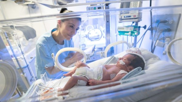 Neonatal nurse checks on a newborn in an incubator with tubes and sensors.