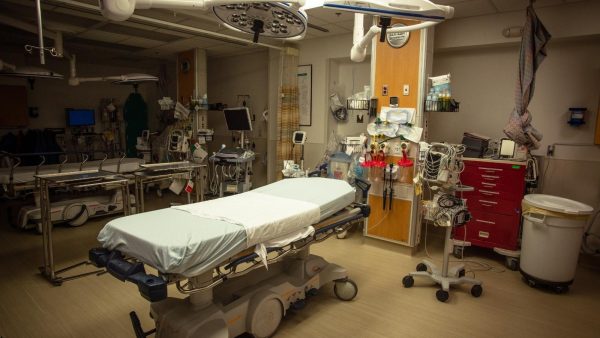 Operating room with an empty patient bed, overhead lights, and numerous medical carts and monitors surrounding it.