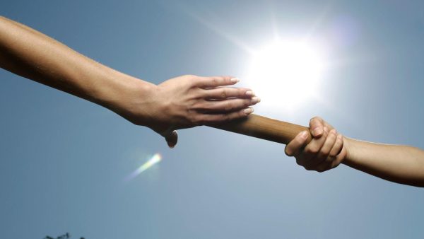 Two people exchange a wooden baton during a relay against a bright sunny sky.