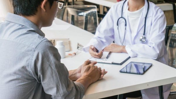 Doctor in white coat and stethoscope consults a patient across a white table, with a clipboard and tablet on the desk.
