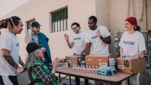 Volunteer group in white shirts labeled VOLUNTEER distribute food and aid from labeled boxes to an elderly man in a wheelchair at a outdoor table.
