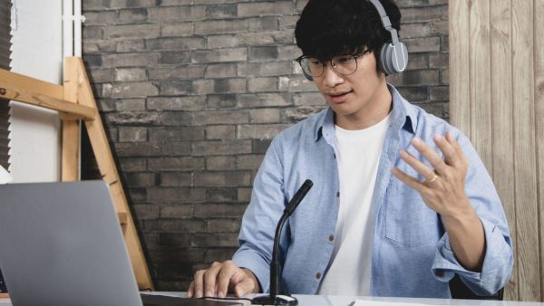 Man wearing headphones and glasses sits at a desk, typing on a laptop and speaking into a desk microphone in a studio setting.