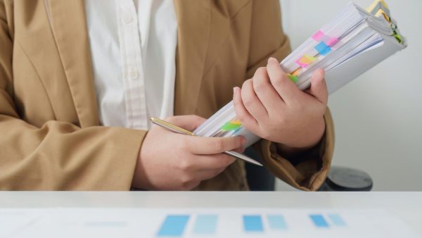 Person in a tan blazer holds a thick stack of documents with colorful sticky tabs while writing with a gold pen.