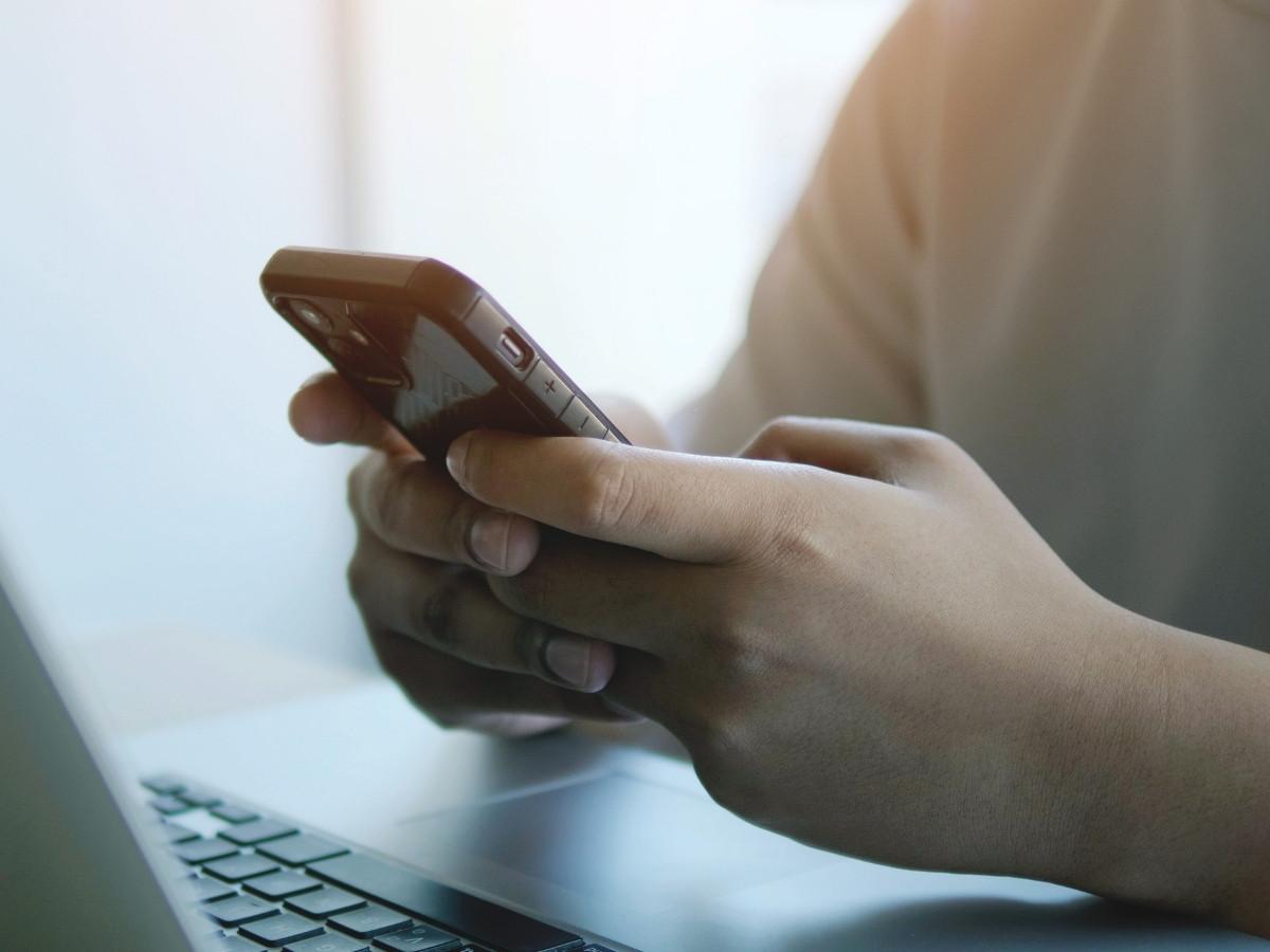 Person holding a smartphone in both hands while a laptop sits nearby, suggesting mobile device use alongside a computer.